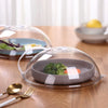 A transparent splash-proof oven cover with a handle sits over a dark gray dish containing green vegetables on a wooden table.