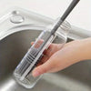 A hand is holding a clear silicone bottle brush with a long handle inside a stainless steel sink for cleaning.