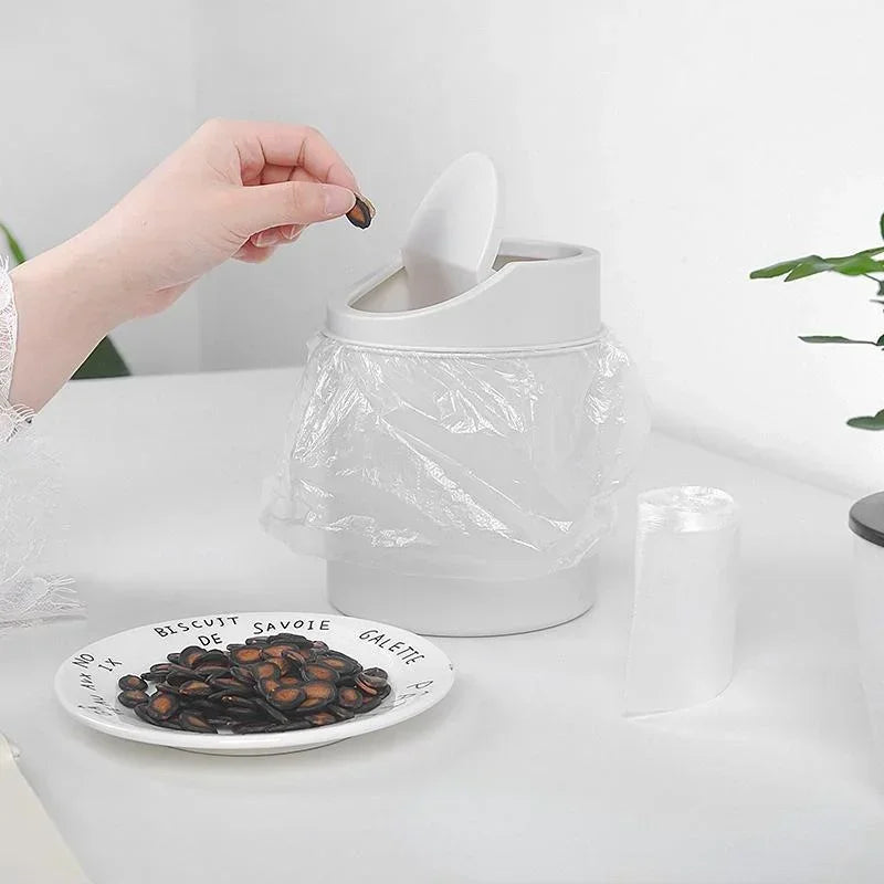 A hand is using a flip cover on a compact desktop trash can, with a plate of biscuits nearby.