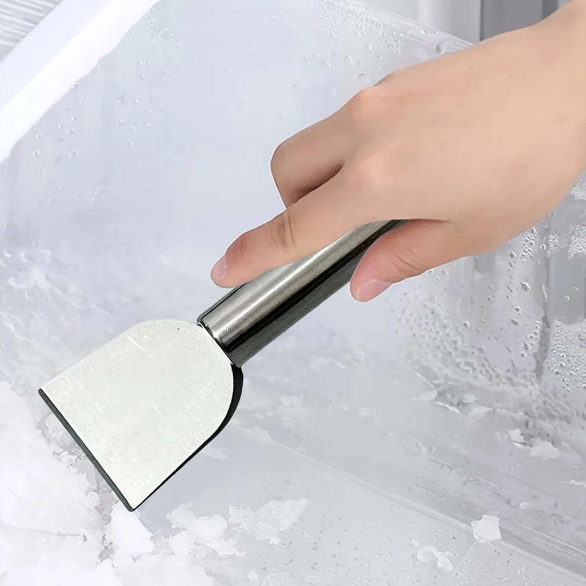 A hand is using a stainless steel multi-purpose scraper to remove snow and ice from a window pane.