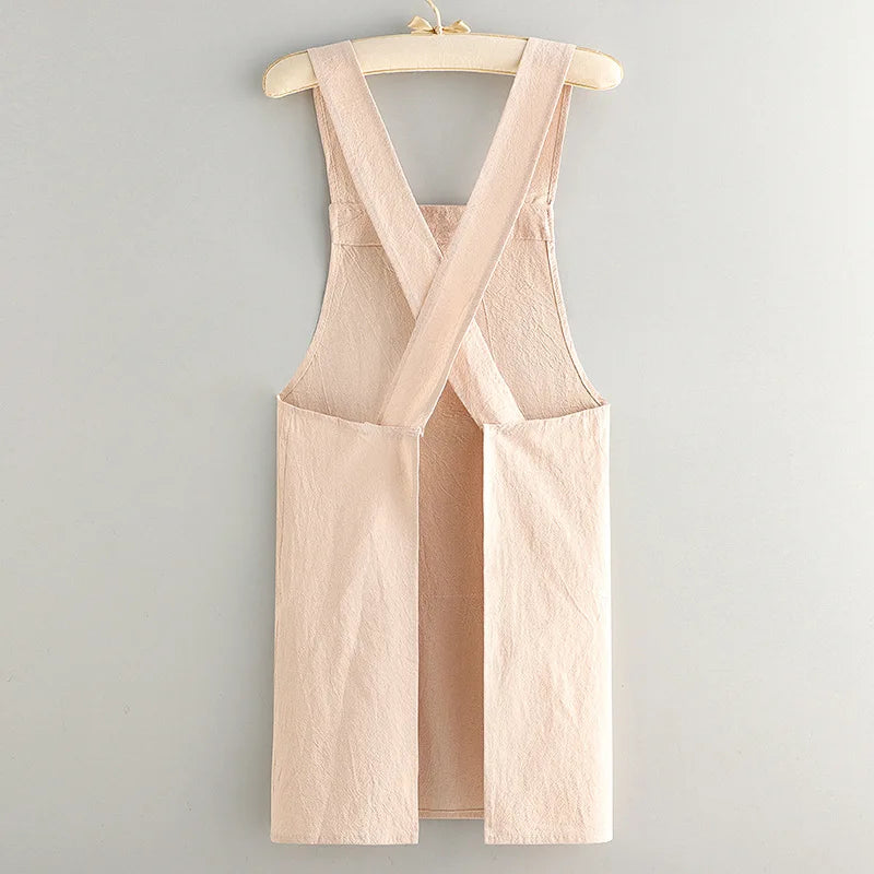 A light beige cotton kitchen apron with criss-cross straps hanging on a wooden hanger.