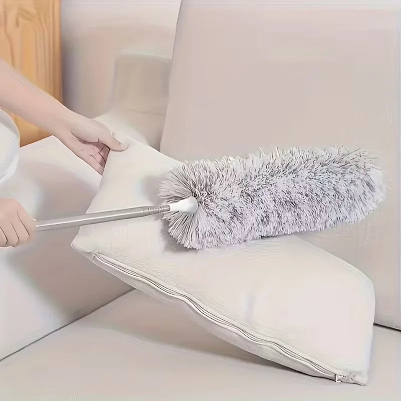 A hand is using a retractable microfiber duster with an extension rod to clean a white pillow.