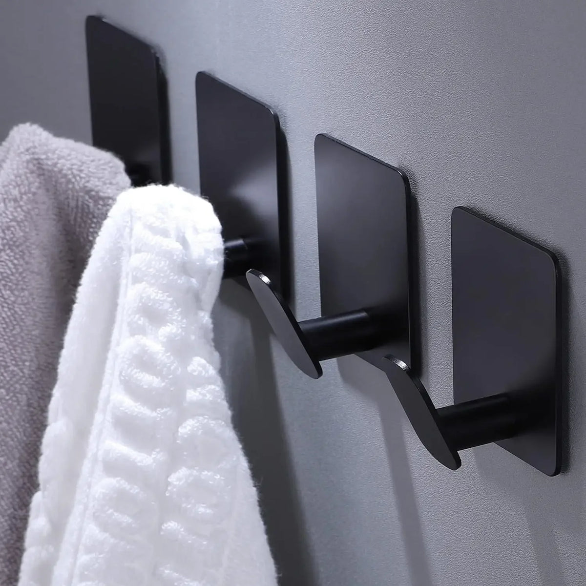 Black stainless steel adhesive hooks mounted on a gray wall, used for hanging towels or kitchen items.