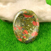 A polished, oval natural quartz thumb stone with green and red inclusions rests on bright green grass.