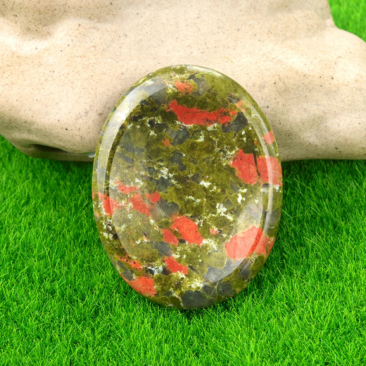 A polished, oval natural quartz thumb stone with green and red inclusions rests on bright green grass.
