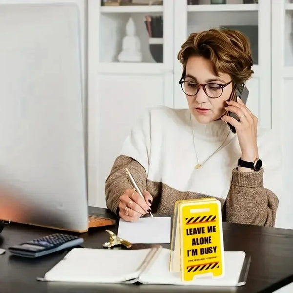 A woman in glasses is holding a phone while sitting at a desk with an acrylic sign that reads Leave Me Alone I'm Busy