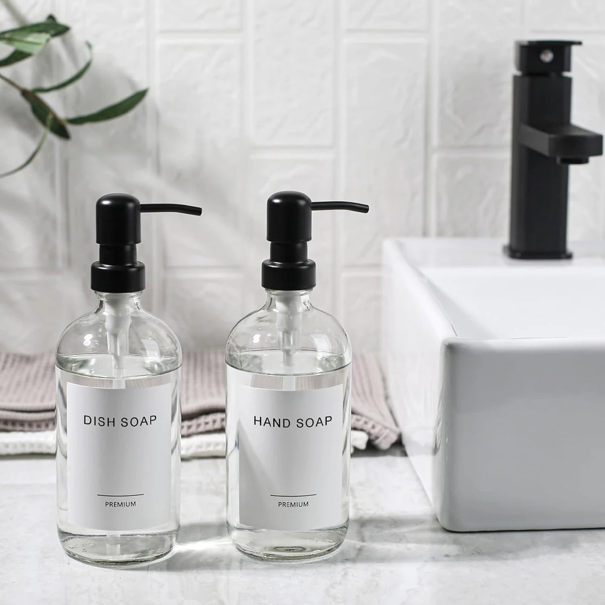 A set of clear glass soap dispensers with black pumps for dish soap and hand soap on a white countertop.