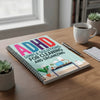 A notebook titled ADHD Simple Solutions for Cleaning and Organizing lies on a wooden table next to a pen and a mug.