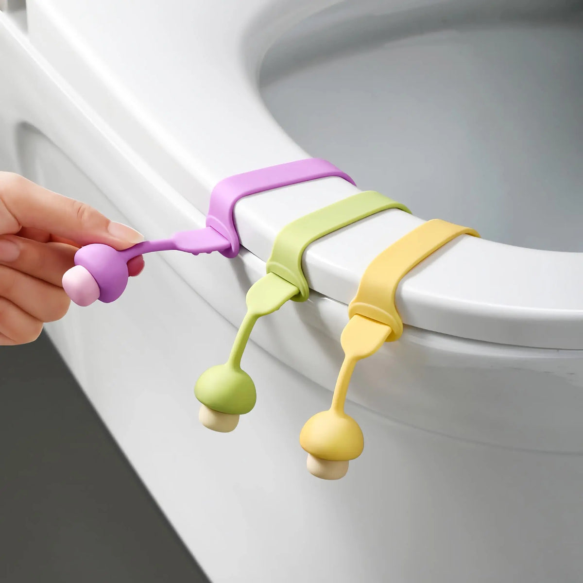A hand is lifting silicone toilet lid lifters in various pastel colors, including purple, green, and yellow.