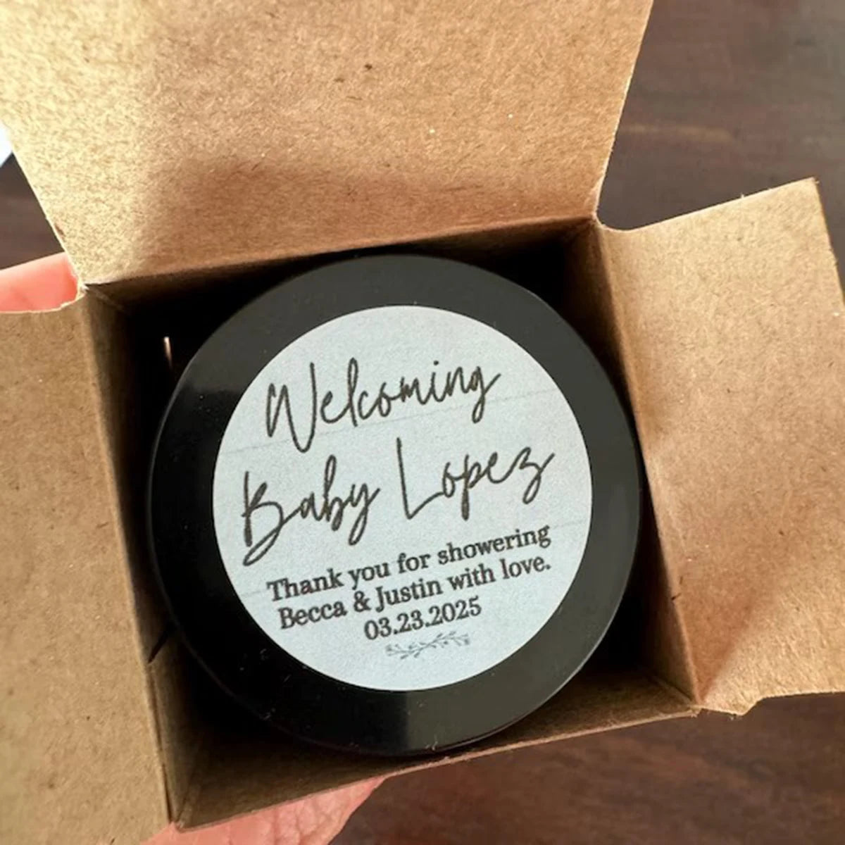 A black round candle jar with white text welcoming Baby Lopez and a thank you message for showering Becca & Justin with love,