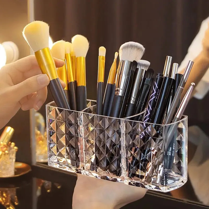 A hand holds a clear acrylic makeup brush holder with three compartments filled with various makeup brushes.