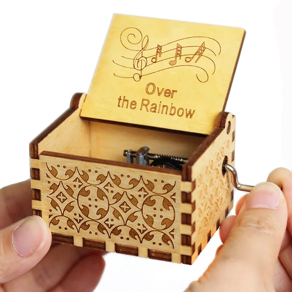 An antique wooden hand crank music box with a lid featuring musical notes and the text Over the Rainbow is held in a hand.