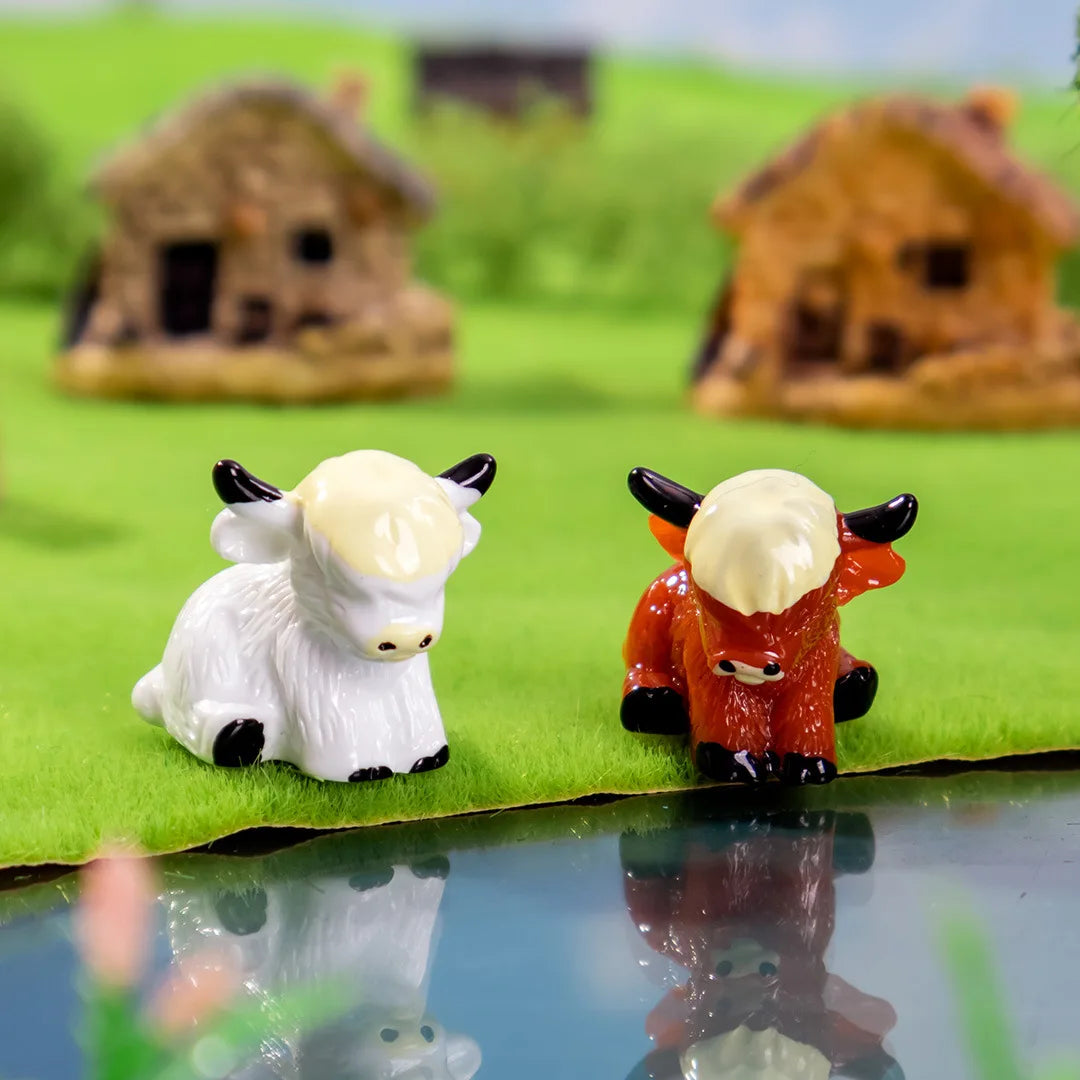 A white resin figurine of a Highland cow with black horns and orange face sits on green grass.