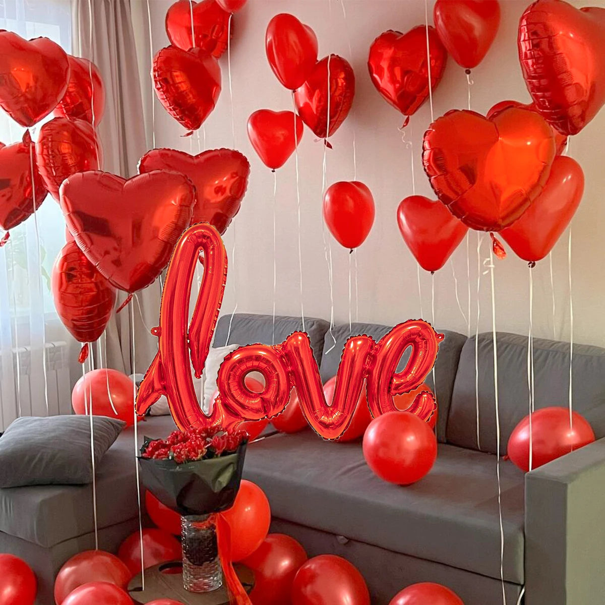 Eine Anordnung roter herzförmiger Ballons und ein großes rotes 'Liebe'-Ballon vor einem grauen Sofa.