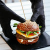 Black gloved hands are assembling a hamburger on a black slate board.