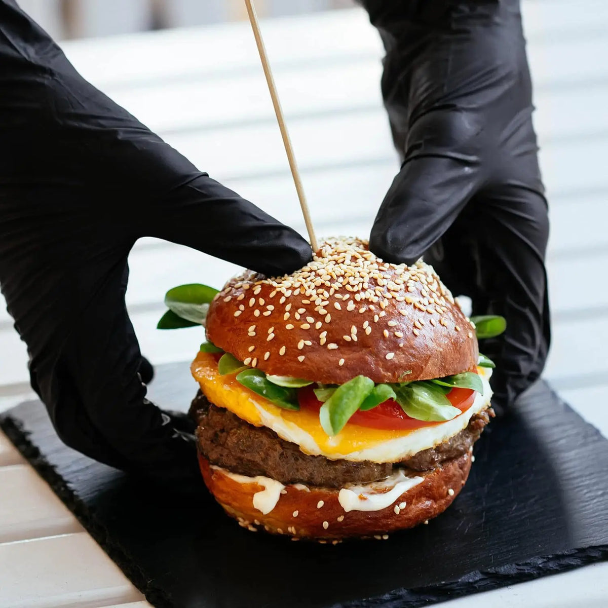 Black gloved hands are assembling a hamburger on a black slate board.