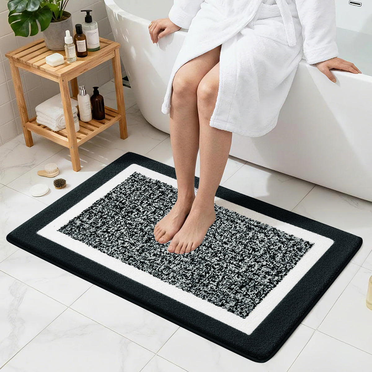 A white bathrobe is shown sitting on a bathtub edge, with a black and white microfiber bath mat placed on the floor.