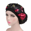 A black satin sleep bonnet with a floral print featuring pink and green leaves, displayed on a mannequin head.