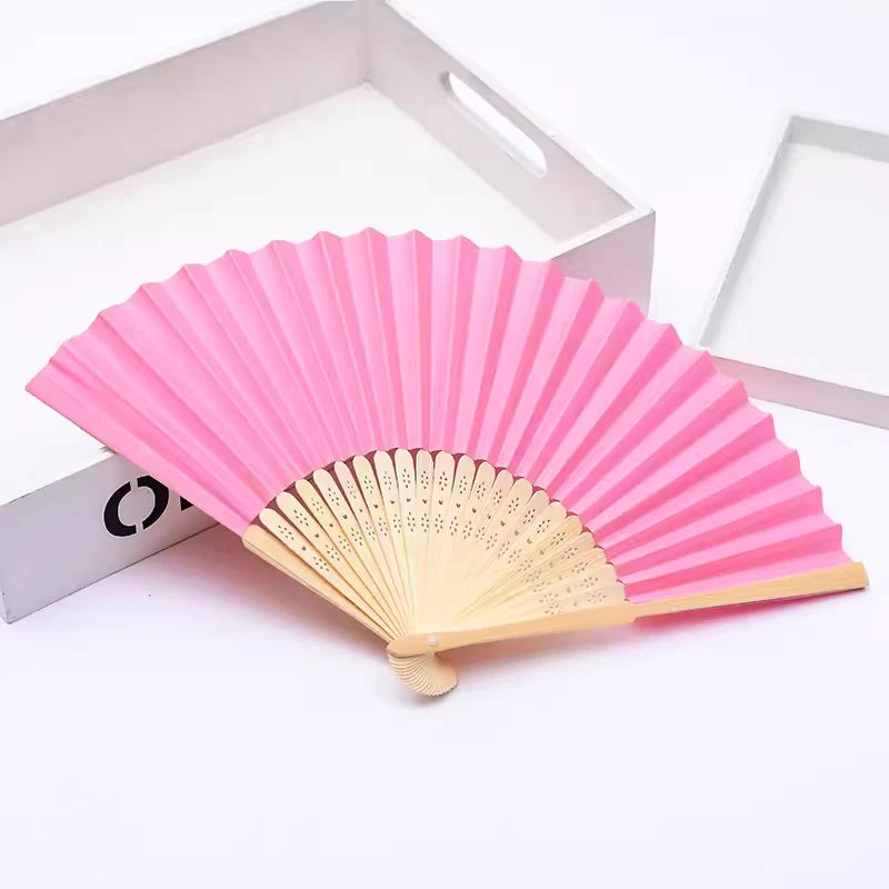 Ventilateur pliable rose en bambou et papier posé dans une boîte blanche.