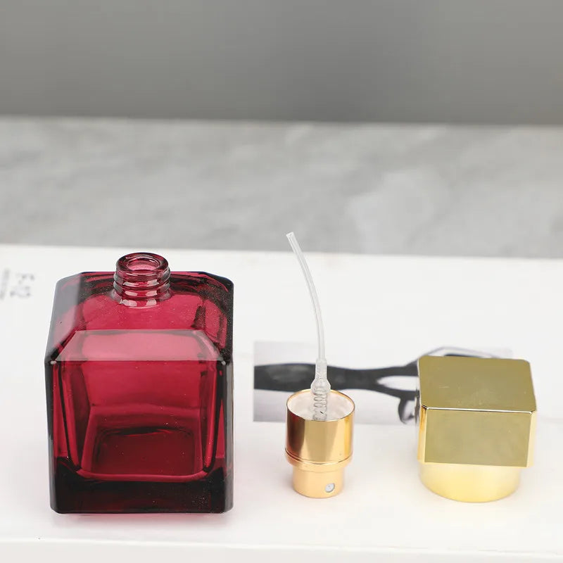 A dark red glass perfume spray bottle next to a gold-colored metal applicator and cap.