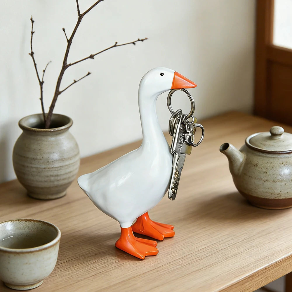 A white goose sculpture with orange feet and a silver key chain attached, displayed on a wooden table.
