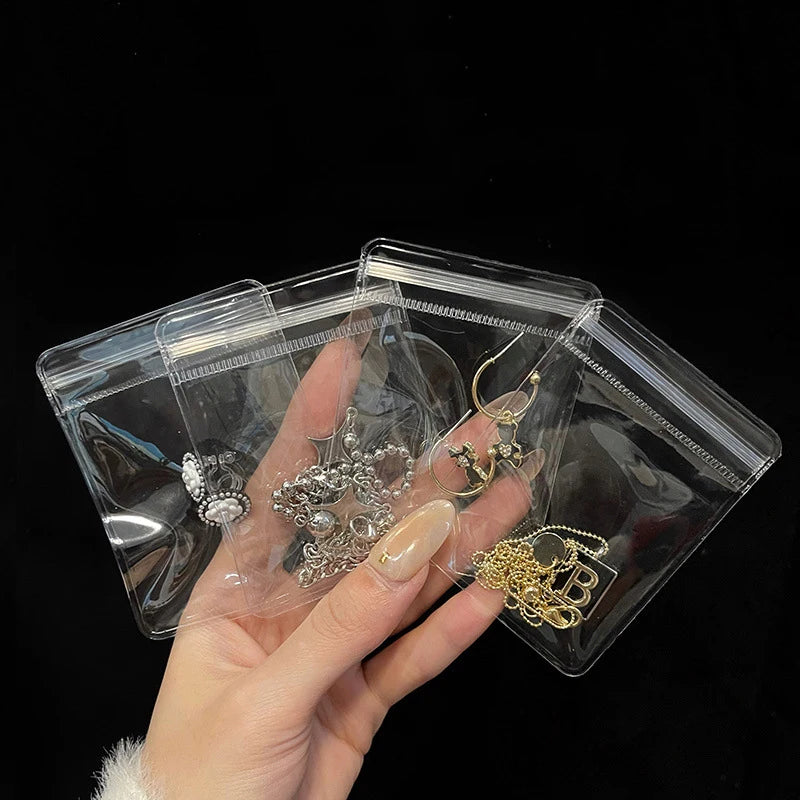 A hand holds clear PVC zip lock jewelry pouches containing various gold and silver necklaces and earrings.