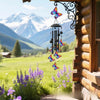 A handcrafted metal wind chime decoration hangs by a window in a rustic wooden cabin with a snowy mountain landscape in the b
