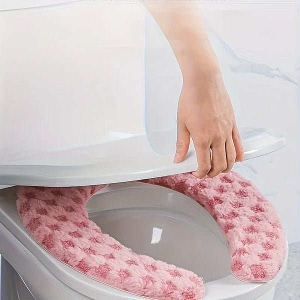 A hand is placing a pink, fluffy washable toilet seat cushion cover onto a white toilet bowl.