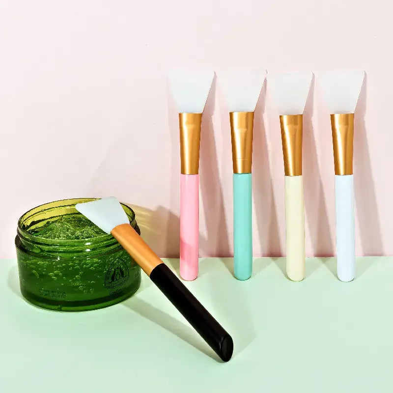 A collection of six silicone facial mask brushes with gold and pastel colored handles displayed on a light surface.
