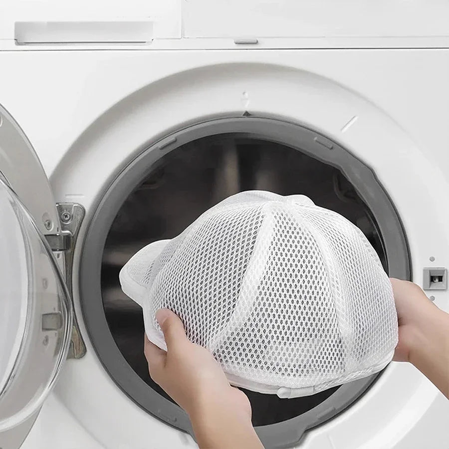 Hands place a white mesh laundry bag over a hat inside a washing machine drum.