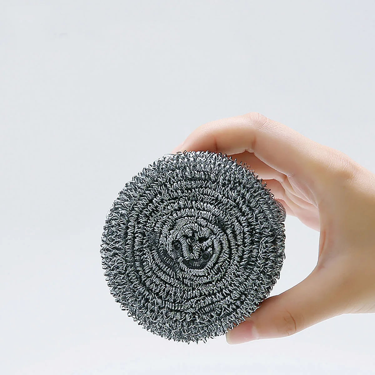 A hand holds a circular pad of gray stainless steel scrubber material against a white background.