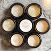 A collection of nine small jars filled with white wax candles arranged in a circular pattern on a light gray surface.