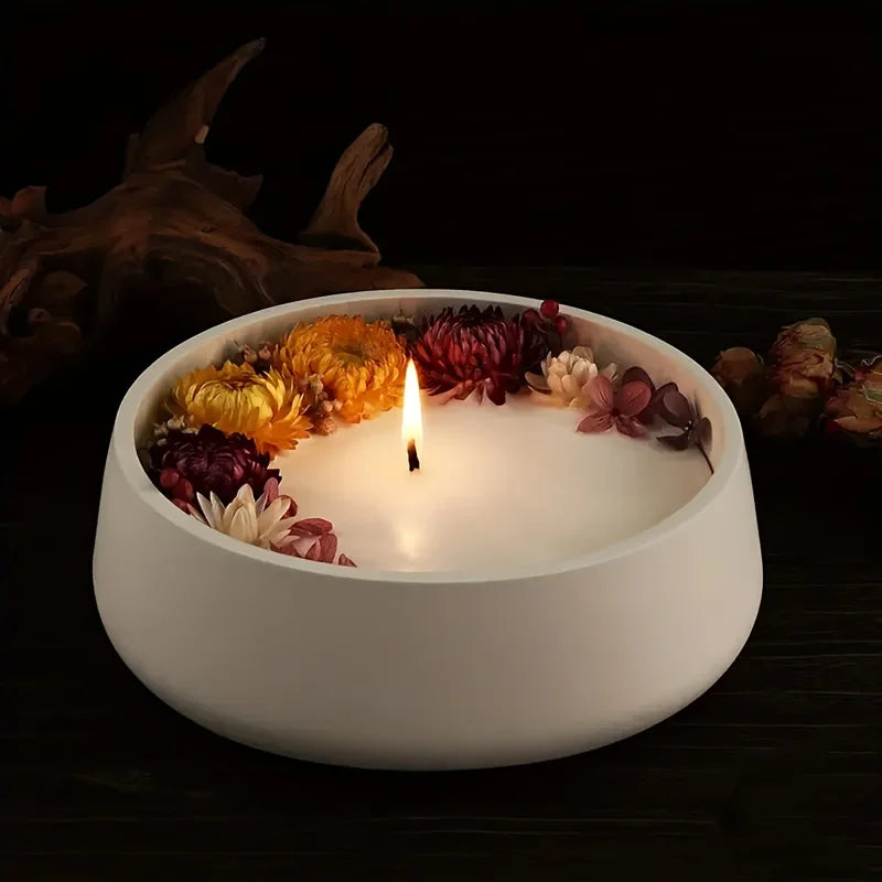 A white candle in a bowl surrounded by dried flowers and a dark wood background.