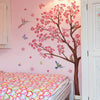 A pink bedroom wall features a large decorative branch design resembling a peach tree with pink blossoms and small butterflie