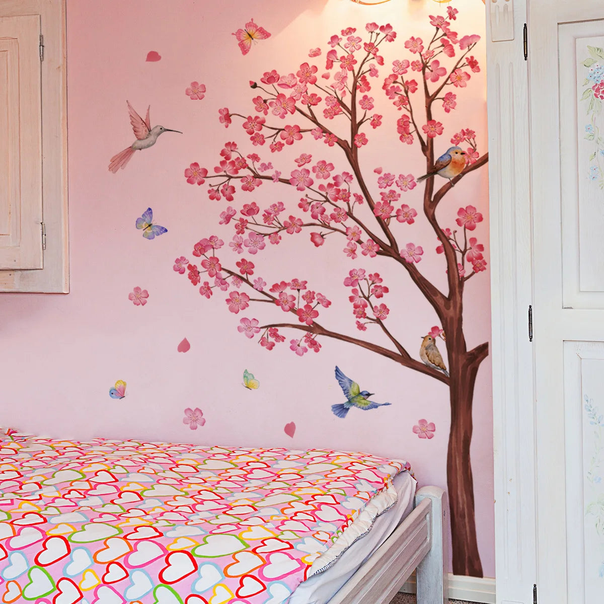 A pink bedroom wall features a large decorative branch design resembling a peach tree with pink blossoms and small butterflie