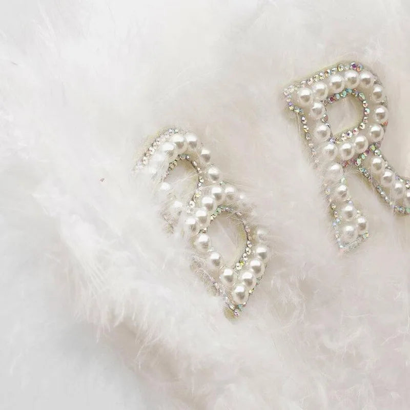 A sparkling pearl initial letter R made of beads is displayed on soft white feathers.