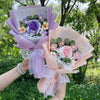 A handmade crochet bouquet featuring purple and pink flowers wrapped in sheer lilac paper and ribbon.