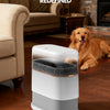 A white, modern slim hands-free trash can with a dark lid sits on a wooden floor next to a golden retriever.