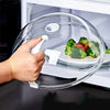 A hand is lifting a clear, splash-proof oven cover with a white handle over a plate of vegetables.