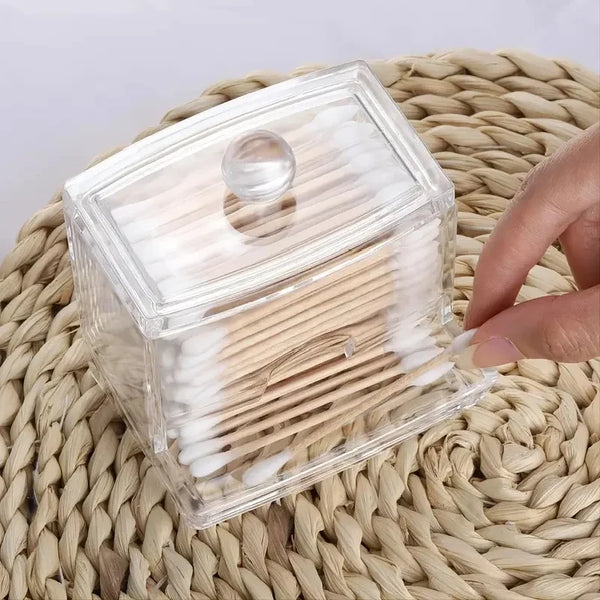 A clear plastic storage box with a lid holds numerous cotton swabs and pads, resting on a woven mat.