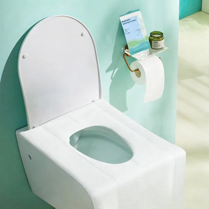 A white bulk disposable toilet seat cover is mounted on a light blue wall, with a roll of toilet paper nearby.