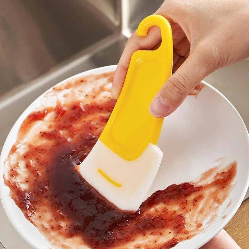 A hand uses a yellow silicone kitchen scraper to clean a white plate covered in red food residue.