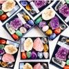 A collection of various natural crystals including amethyst, rose quartz, citrine, and green stones arranged in black trays.
