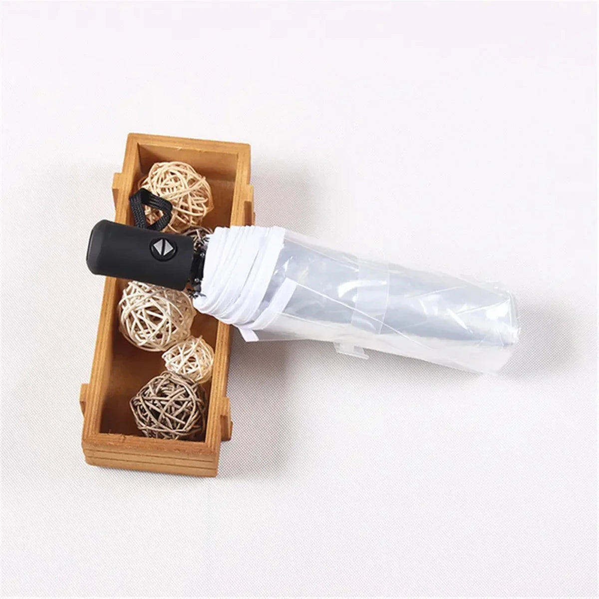 A black automatic transparent folding umbrella is shown next to a wooden box filled with natural straw balls.