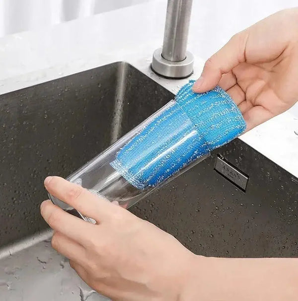 A hand is holding a blue double-sided non-scratch dishwashing sponge set over a kitchen sink.