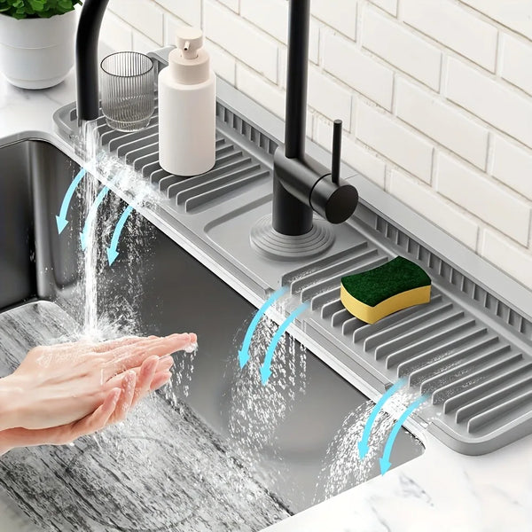 A hand is washing under a silicone kitchen faucet splash guard mat with water spraying onto the counter.