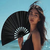 A woman holds a black handheld folding fan open against a blue sky and ocean backdrop.