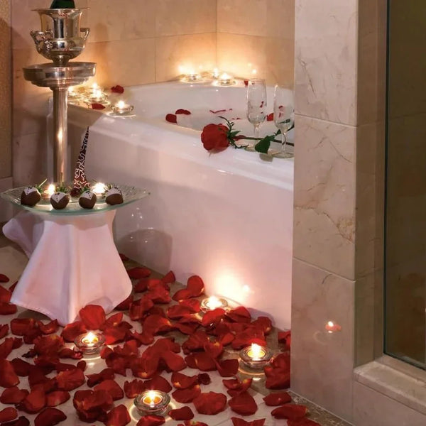 Red artificial silk rose petals scattered on the floor around a white bathtub with lit candles.