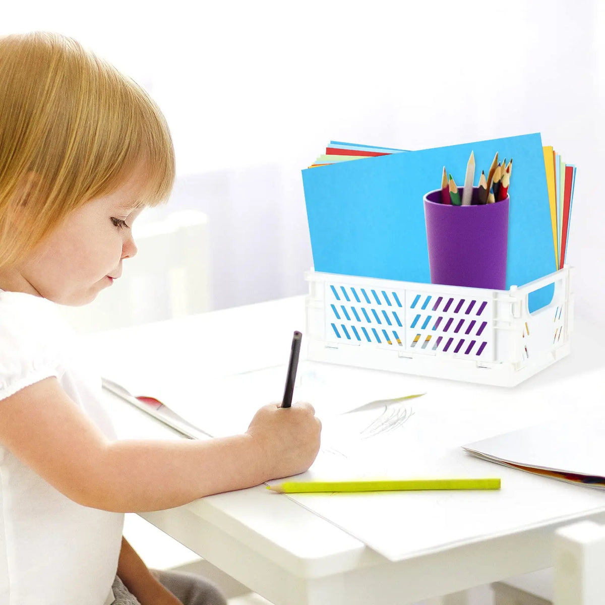 A young girl is writing at a table next to a purple storage basket filled with colored pencils and paper.