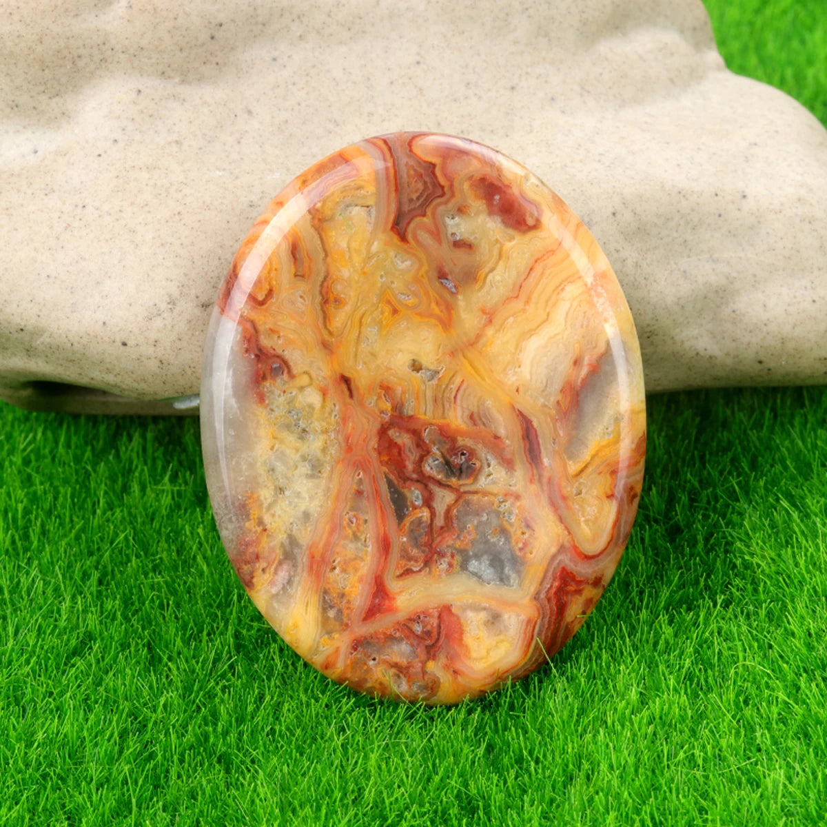 A polished, oval piece of natural quartz stone with swirling shades of orange, brown, and amber resting on green grass.